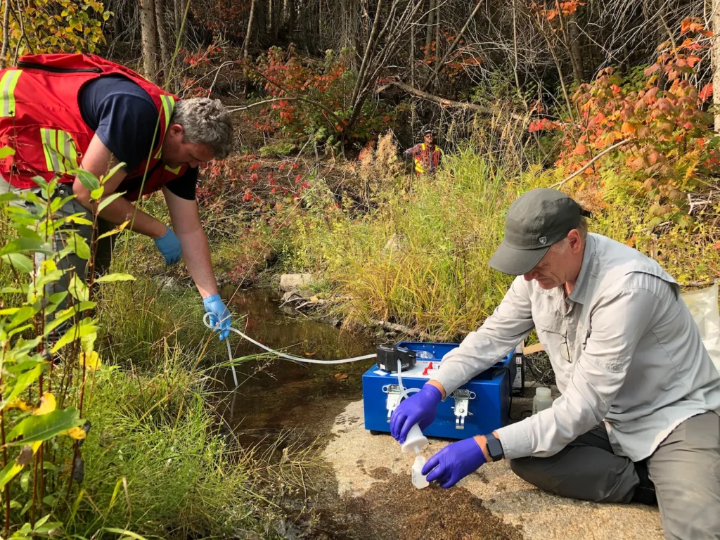 Two geochemists collecting a filtered stream water sample beside a woodland creek in autumn