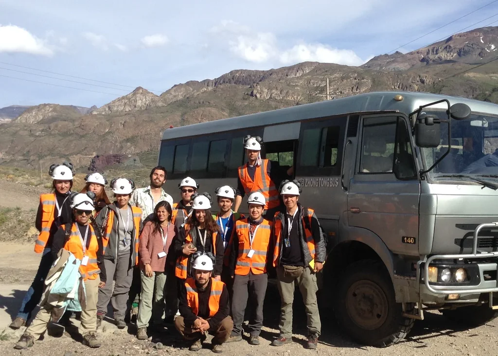 Students and geologists in hard hats and high-visibility vests beside a field bus in mountain terrain